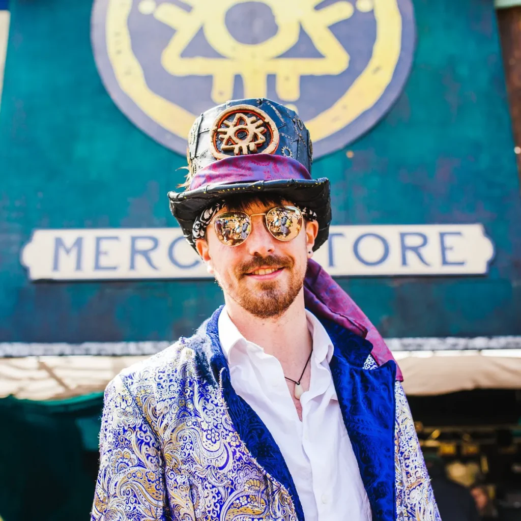 Festival-goer in a blue and gold brocade tailcoat and steampunk hat with mirrored sunglasses at Boomtown Fair.