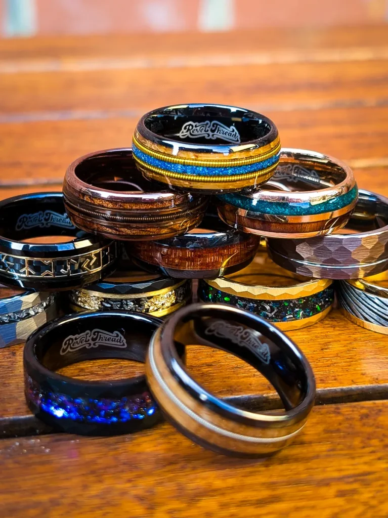 A vibrant stack of handcrafted Revel Threads rings featuring wood, opal, guitar string, and metal inlays on a wooden table.