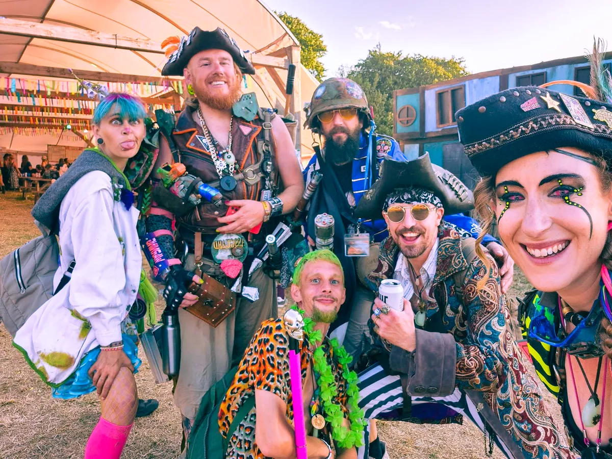 Group of festival-goers in colorful, elaborate costumes at Boomtown Fair, smiling and posing under a marquee.
